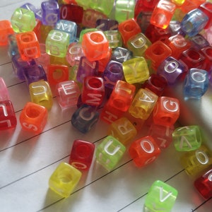 Può includere: Un mucchio di perline di lettere in plastica colorate, con lettere come A, B, C, D, E, F, G, H, I, J, K, L, M, N, O, P, Q, R, S, T, U, V, W, X, Y e Z. Le perline sono traslucide e hanno lettere bianche.