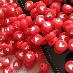 May include: A close-up of a large pile of red heart-shaped beads with white hearts in the centre. The beads are scattered on a black surface.