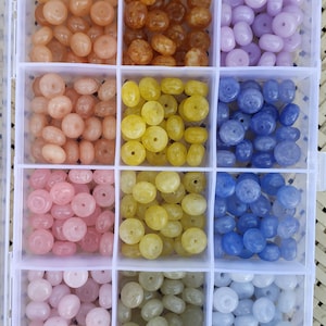 May include: A clear plastic organizer filled with beads in various colors. The beads are in shades of peach, brown, lavender, pink, yellow, and blue. The beads are round with a hole through the center, perfect for jewelry making.
