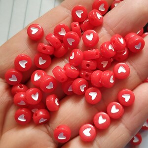 May include: A collection of red heart-shaped beads with white hearts in the centre. The beads are scattered on a hand.