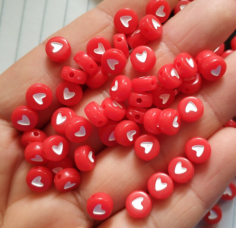May include: A collection of red heart-shaped beads with white hearts in the centre. The beads are scattered on a hand.