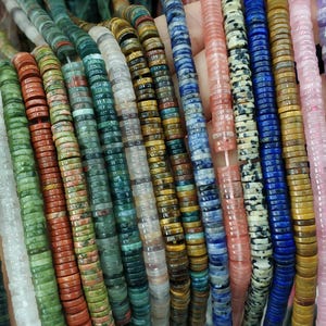 May include: A selection of colourful gemstone beads in shades of green, red, blue, and pink. The flat, round beads are strung together, displaying diverse textures and patterns. Suitable for jewellery making.