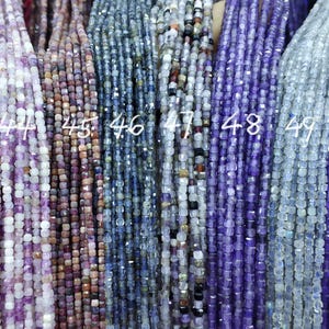 Può includere: Primo piano di più fili di perline di pietre preziose in vari colori, tra cui sfumature di viola, blu e rosa. Le perline sono piccole e di forma quadrata, infilate insieme per creare un aspetto strutturato. I numeri sono visibili.