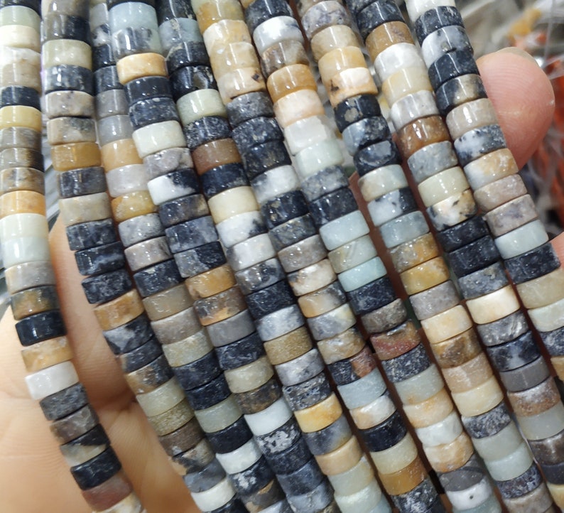 May include: A strand of multi-colored gemstone beads. The beads are round and have a mottled appearance, with shades of brown, black, white, and green. The beads are strung on a clear thread.