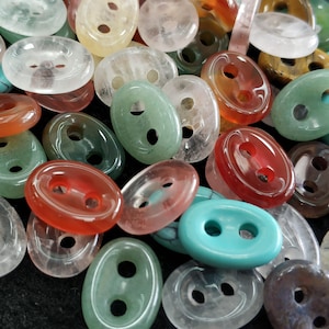 Może przedstawiać: Kolekcja owalnych guzików w różnych kolorach, w tym przezroczystym, czerwonym, zielonym i turkusowym. Każdy guzik ma dwa otwory do szycia. Guziki wykonane są z półprzezroczystego materiału.