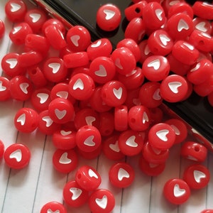 May include: A pile of red plastic beads with white heart designs. The beads are scattered on a white surface with black lines.