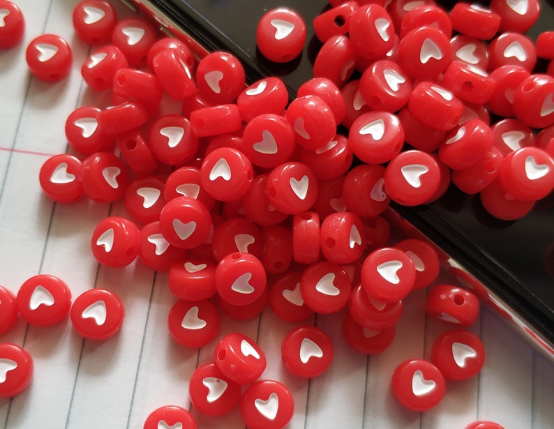 May include: A pile of red plastic beads with white heart designs. The beads are scattered on a white surface with black lines.