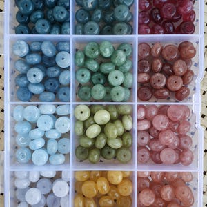 Op de afbeelding: Een doorzichtige plastic organizer gevuld met edelsteen kralen in verschillende kleuren. De kralen zijn in tinten blauw, groen, rood, bruin, geel en roze. Elk compartiment bevat een andere kleur ronde, donutvormige kralen, perfect voor het maken van sieraden.
