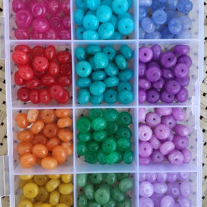 Puede incluir: Un organizador de plástico transparente lleno de cuentas de colores. Las cuentas son de varios tonos de rosa, azul, rojo, verde, naranja, amarillo y morado. Cada color está separado en su propio compartimento, ofreciendo una amplia variedad para la fabricación de joyas y manualidades.