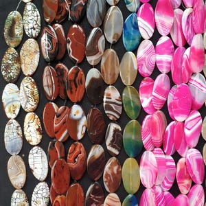 May include: A collection of oval-shaped gemstone beads in various colors and patterns. The beads are strung together and include shades of brown, red, white, gray, pink, and blue. The gemstones have unique veining and marbling.