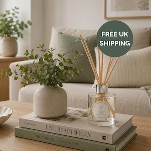 May include: A clear glass reed diffuser with a silver collar and wooden reeds, featuring a circular label that reads "HOME AT NUMBER TWENTY TWO" and "ROSE". The diffuser sits on two stacked books titled "LIVE BEAUTIFULLY" and "THIS IS HOME".