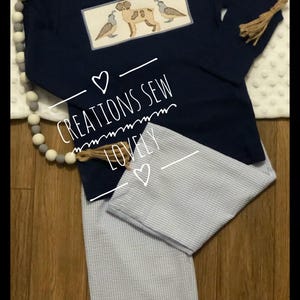 May include: A navy blue long-sleeved shirt with a white embroidered patch featuring a dog and two birds. The shirt has a white and blue striped pants with a brown tassel. The text "CREATIONS SEW LOVELY" is written on the shirt.