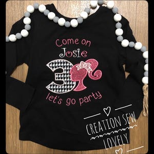 May include: Black long-sleeved shirt with a pink and white embroidered design featuring a Barbie doll and the number 3. The text "Come on Josie let's go party" is also embroidered on the shirt.
