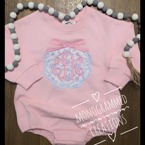 May include: Pink baby bodysuit with long sleeves, featuring a floral monogram design in blue and pink. The bodysuit has a bow detail at the top and the text "MONOGRAMMED CREATIONS".