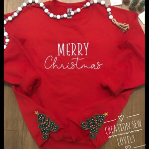 May include: A red Christmas sweatshirt with the words "MERRY Christmas" in white script. The sleeves feature green Christmas tree appliqués with white dots and gold star accents. A decorative garland with gray and white beads is draped across the top.