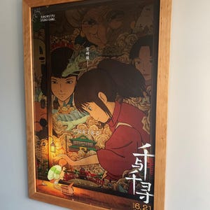 May include: Framed poster of the animated film "Spirited Away." The poster is in a wooden frame and shows a young girl in a red outfit, with Japanese text and imagery of fantastical creatures. The poster is mounted on a light blue wall.