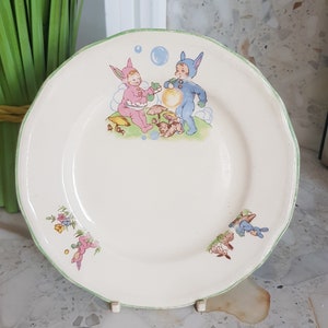 May include: A vintage white ceramic plate with a green rim features a whimsical illustration of two children dressed as bunnies, one in pink and one in blue, playing with bubbles. The plate is decorated with additional bunny illustrations and floral accents.