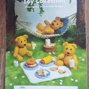 May include: A knitting pattern booklet titled Jean Greenhowe's Toy Collection featuring over twenty easy-to-knit designs. The cover displays three yellow knitted teddy bears having a picnic with knitted food items on a white blanket.