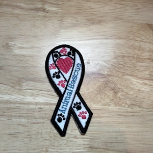 Peut inclure: Écusson de ruban brodé avec les mots "Animal Rescue" en bleu. Le patch présente un cœur rouge et des empreintes de pattes en noir et rouge. Le ruban a une bordure noire et est sur une surface en bois.
