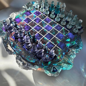 May include: A unique, handmade chess set with a shimmering, iridescent resin board and clear, blue, and purple chess pieces. The board features a grid pattern with a galaxy-like design, and the pieces are arranged for play.