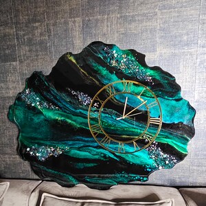 May include: A unique wall clock with a gold-colored frame and Roman numerals. The clock face is set against a black and teal resin background with glitter accents. The clock has a freeform, organic shape.