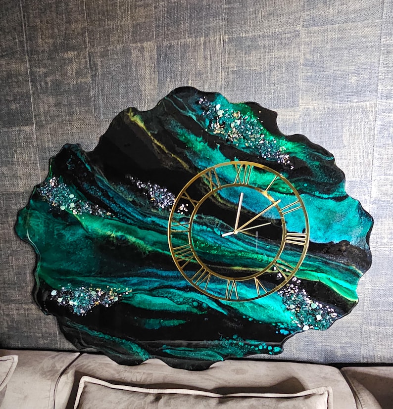 May include: A unique wall clock with a gold-colored frame and Roman numerals. The clock face is set against a black and teal resin background with glitter accents. The clock has a freeform, organic shape.