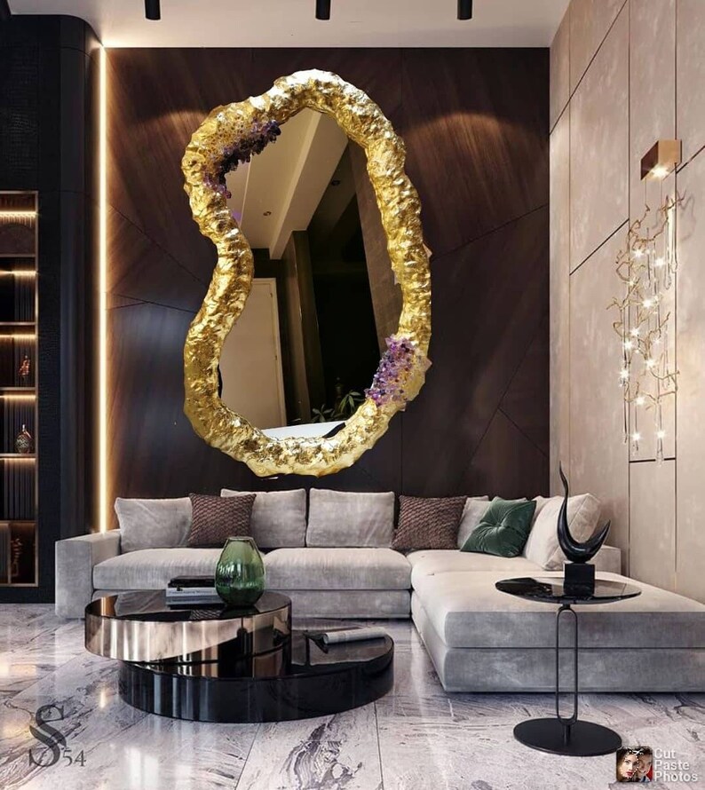 May include: A large, irregularly shaped gold mirror with a textured surface hangs on a dark wood wall. The mirror is adorned with purple floral accents. The room features a gray sectional sofa and modern decor.