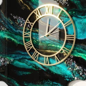 May include: A wall clock with a gold-colored frame and Roman numerals. The clock face is transparent, set against a swirling background of black, teal, and green resin. Decorative accents are scattered across the surface.