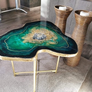 Könnte beinhalten: Einzigartiger, handgefertigter Couchtisch mit goldfarbenem Metallgestell. Die Tischplatte hat ein auffälliges Design mit Grün-, Gold- und Schwarztönen, das einer Achatscheibe ähnelt. Zwei hohe, gewebte braune Kerzenhalter sind im Hintergrund.