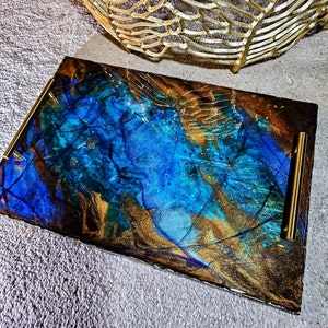 Puede incluir: Una bandeja rectangular para servir con asas doradas. La bandeja está hecha de resina y tiene un diseño marmoleado azul, dorado y marrón.