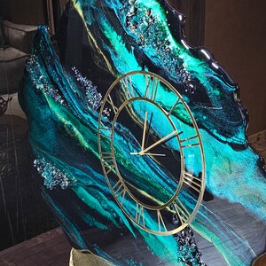 May include: A decorative wall clock with a unique design. The clock face is gold with Roman numerals. The background features a swirling pattern of teal, black, and gold resin, with embedded crystals. The clock is mounted on a gold stand.