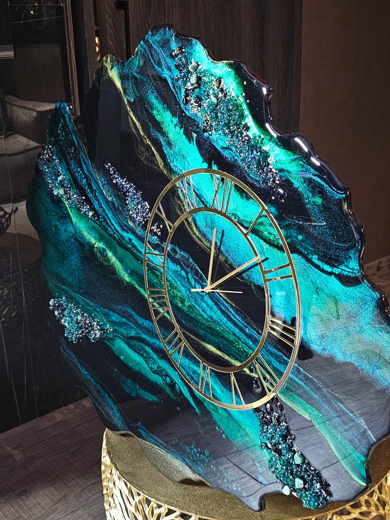 May include: A decorative wall clock with a unique design. The clock face is gold with Roman numerals. The background features a swirling pattern of teal, black, and gold resin, with embedded crystals. The clock is mounted on a gold stand.