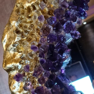 May include: Close-up of a decorative mirror with a gold-colored, textured frame. The frame is adorned with clear and purple crystal-like embellishments, creating a luxurious and artistic design. The mirror's surface reflects the surrounding environment.