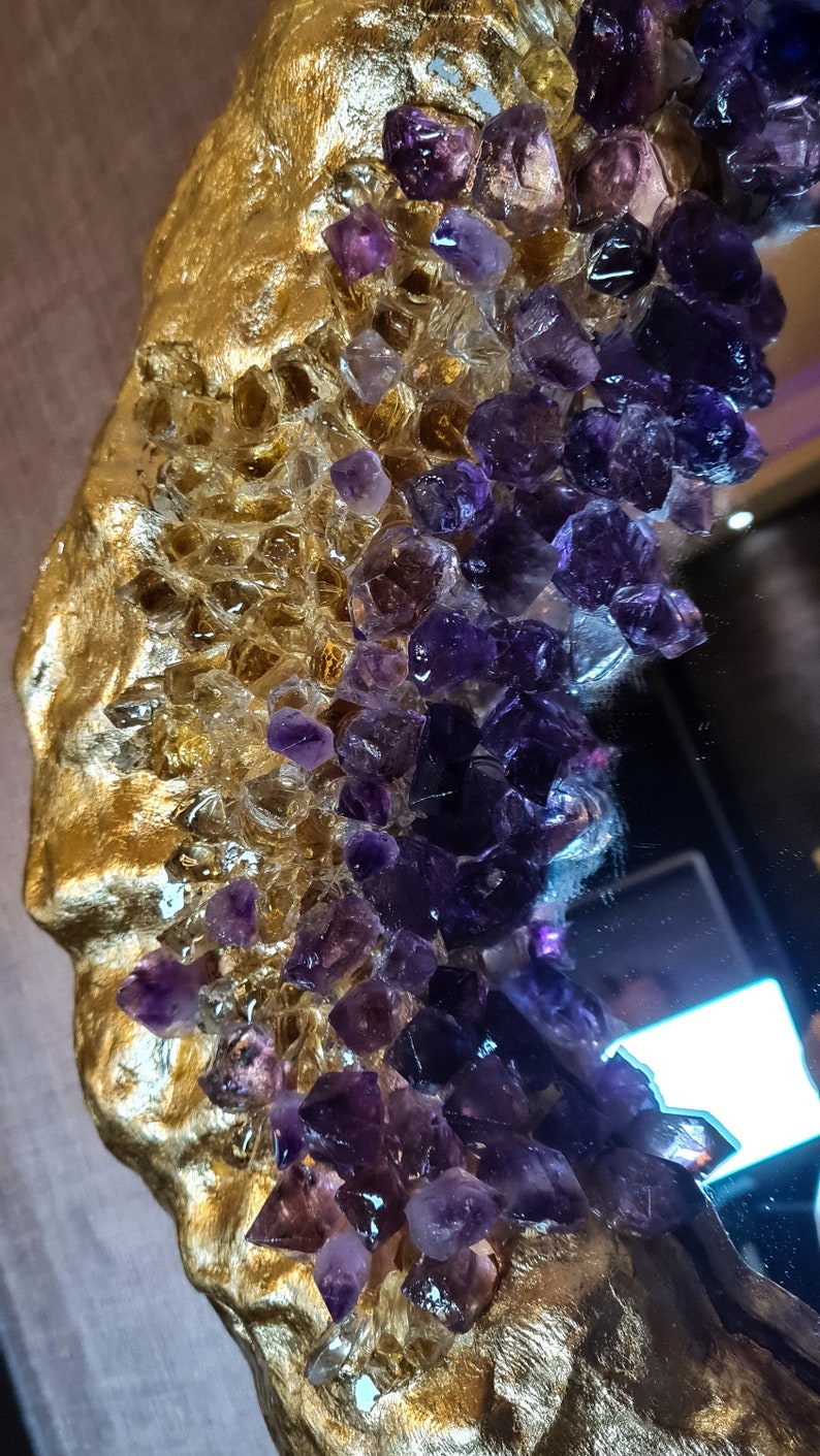 May include: Close-up of a decorative mirror with a gold-colored, textured frame. The frame is adorned with clear and purple crystal-like embellishments, creating a luxurious and artistic design. The mirror's surface reflects the surrounding environment.