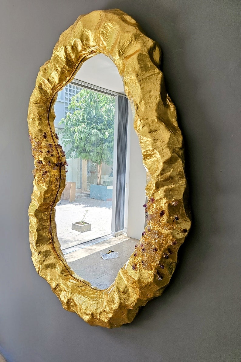 May include: A gold-colored, irregularly shaped mirror with a textured, organic frame. The frame is adorned with small, sparkling crystals. The mirror reflects a view of a bright outdoor scene. A unique decorative piece.