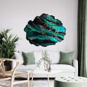 May include: A large, abstract wall clock with a gold-colored frame and Roman numerals. The clock face features a swirling pattern of teal, black, and gold colors, with glitter accents. The clock is mounted on a white wall.