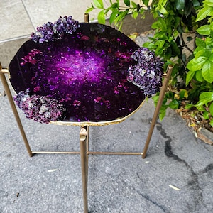 Könnte beinhalten: Ein kleiner, runder Tisch in Lila und Gold mit einer Tischplatte aus Harz und einem goldenen Metallgestell. Die Tischplatte hat ein galaktisches Design mit eingebetteten lila Blumen.