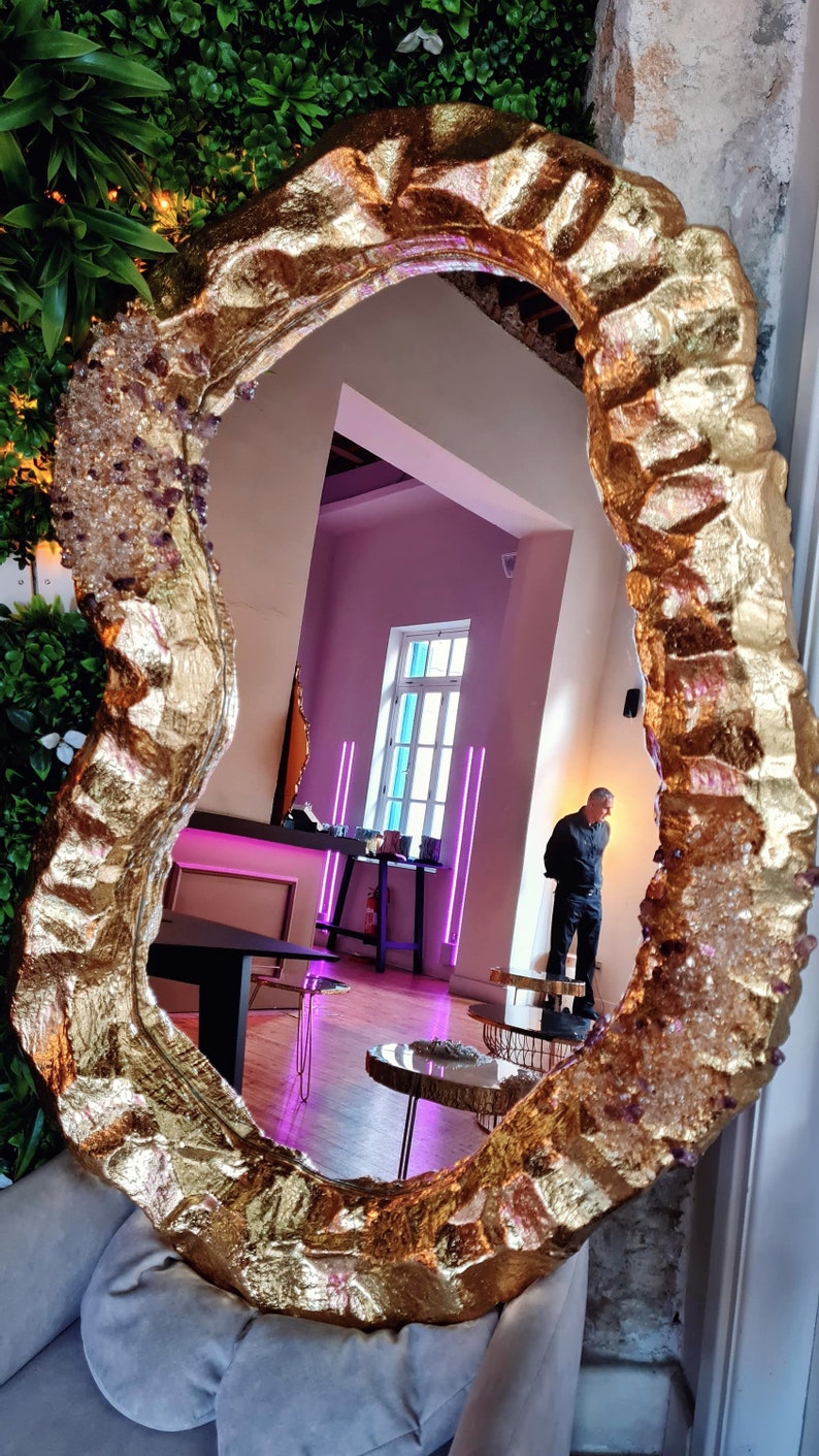 May include: A large, irregularly shaped mirror with a gold-colored, textured frame. The frame is embellished with crystal-like accents. The mirror reflects a room with a person standing near a table and a window with a view.