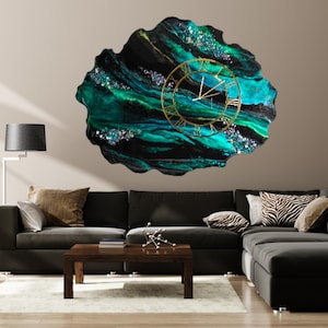 May include: A large, abstract wall clock with a gold-colored frame and Roman numerals. The clock face is set against a swirling background of teal, black, and green resin with glitter accents. The clock is mounted on a neutral-colored wall above a dark gray sectional sofa.