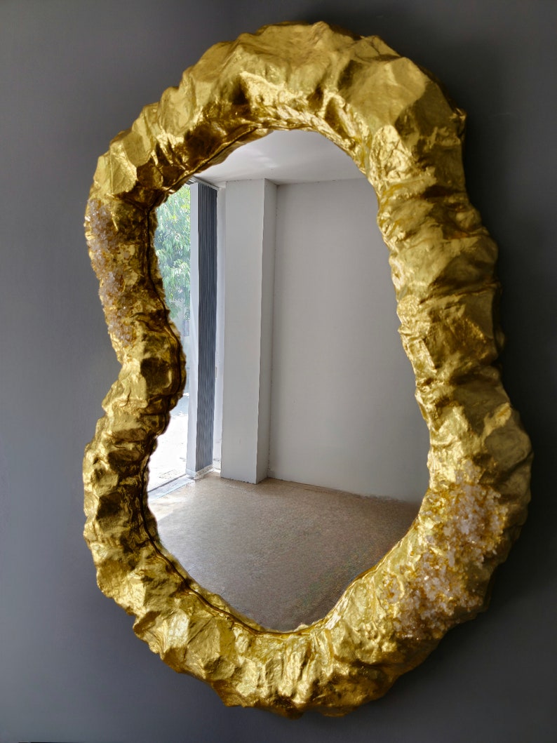 May include: A gold-colored, irregularly shaped wall mirror. The frame has a textured, rough appearance, resembling a rock formation. The mirror reflects a room with a window and a light-colored floor. The overall design is artistic and decorative.