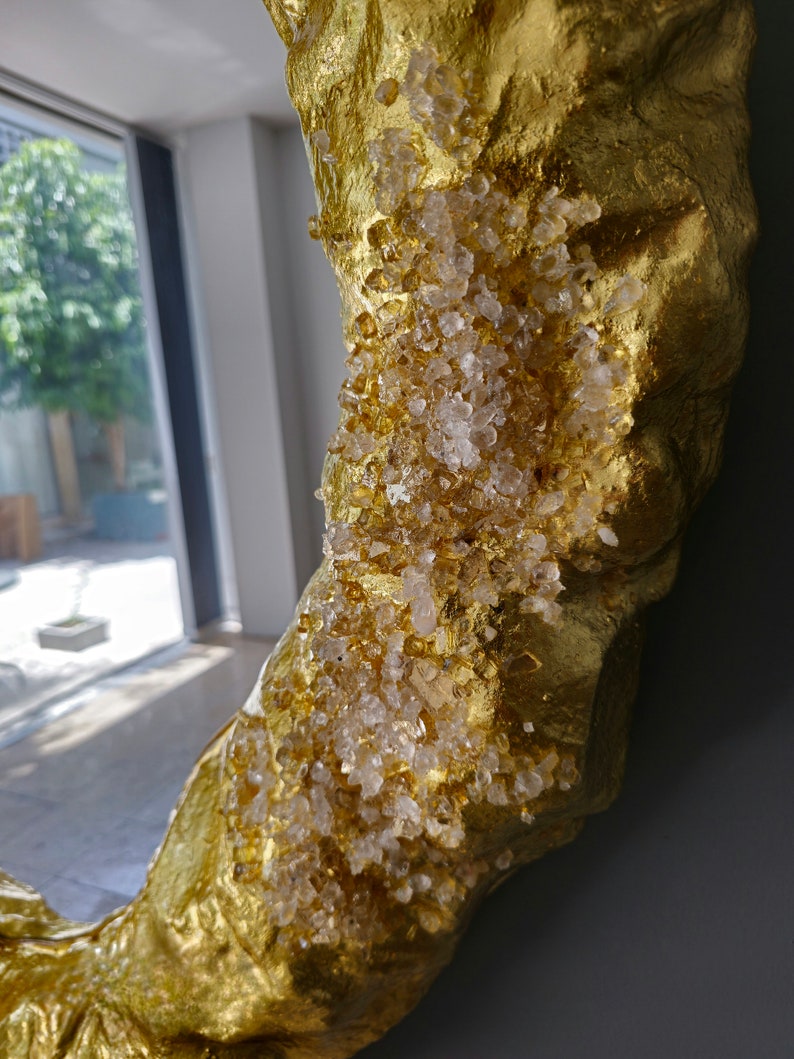 May include: Close-up of a gold-colored, textured decorative object, possibly a mirror frame. The surface is embellished with clear, crystalline accents. The object is set against a gray background, with a glimpse of a window and greenery.