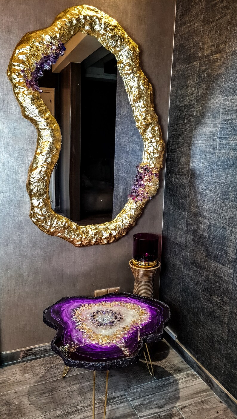 May include: A gold-leafed mirror with an organic shape, adorned with purple and clear crystals. Below, a matching side table features a purple and gold resin top with gold legs. A decorative lamp sits nearby.