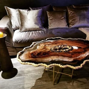 May include: A unique coffee table with a live edge, dark wood top and a gold metal base. The top is shaped like a large, irregular oval with a wavy edge. The wood has a natural, organic look with a dark brown colour and a light brown, almost white, centre. The gold metal base is made of thin, straight bars that form a simple, geometric design.