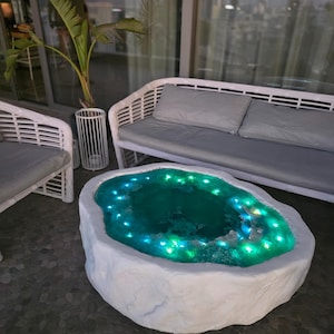 Op de afbeelding: Een unieke, witte salontafel met een steenachtige uitstraling en een centraal, verlicht, teal-kleurig bassin. De tafel wordt omringd door een witte rotan bank met grijze kussens en een potplant, wat een moderne buitenruimte creëert.
