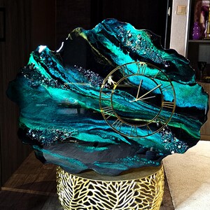 May include: A decorative clock with a unique design. The clock face is gold-colored with Roman numerals. The clock's face is set against a teal, black, and gold resin base. The base sits on a gold-colored, leaf-patterned stand.
