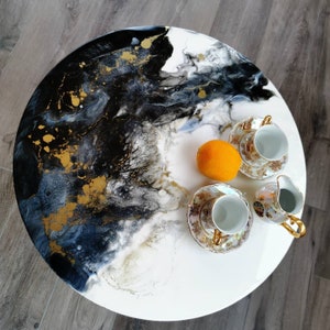 May include: Round coffee table with a white surface, featuring a black, gold, and white abstract design. The table is adorned with two teacups, saucers, a creamer, and an orange. The table's diameter is approximately 30 inches.
