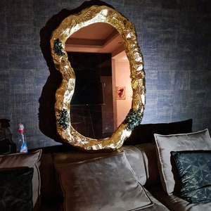 May include: A gold-colored, irregularly shaped mirror with a textured frame. The mirror is mounted on a dark, textured wall. Small clusters of green and black embellishments are visible on the frame. The mirror reflects a room with a painting.