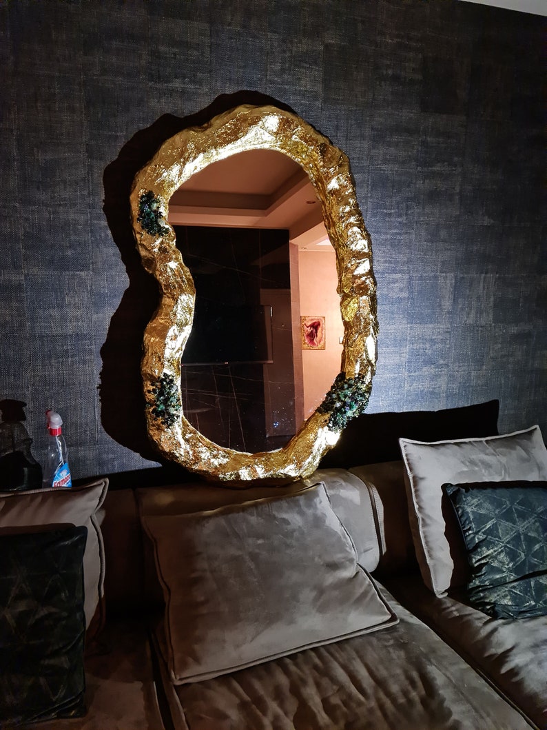 May include: A gold-colored, irregularly shaped mirror with a textured frame. The mirror is mounted on a dark, textured wall. Small clusters of green and black embellishments are visible on the frame. The mirror reflects a room with a painting.