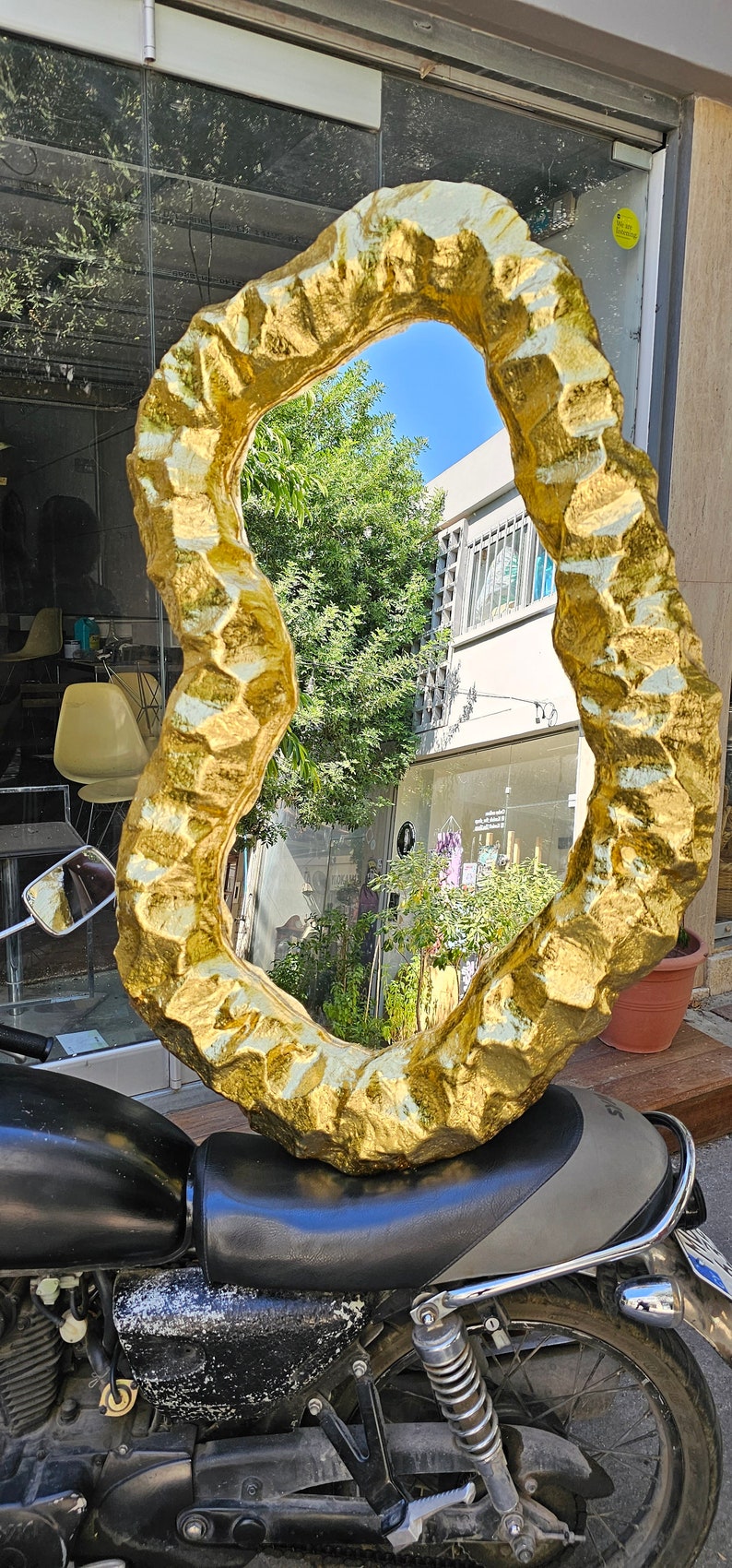 May include: A large, irregularly shaped mirror with a textured, gold-colored frame. The mirror is resting on the seat of a black motorcycle. The background includes a storefront and greenery.