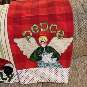 May include: Red and white checkered kitchen towel with a green and white angel holding a white dove. The angel has blonde hair and blue eyes. The towel has the word "peace" embroidered in green on it.